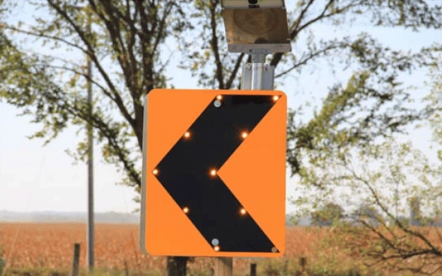 An orange road sign displays a single black chevron with ten miniature lights placed along the chevron's edge.